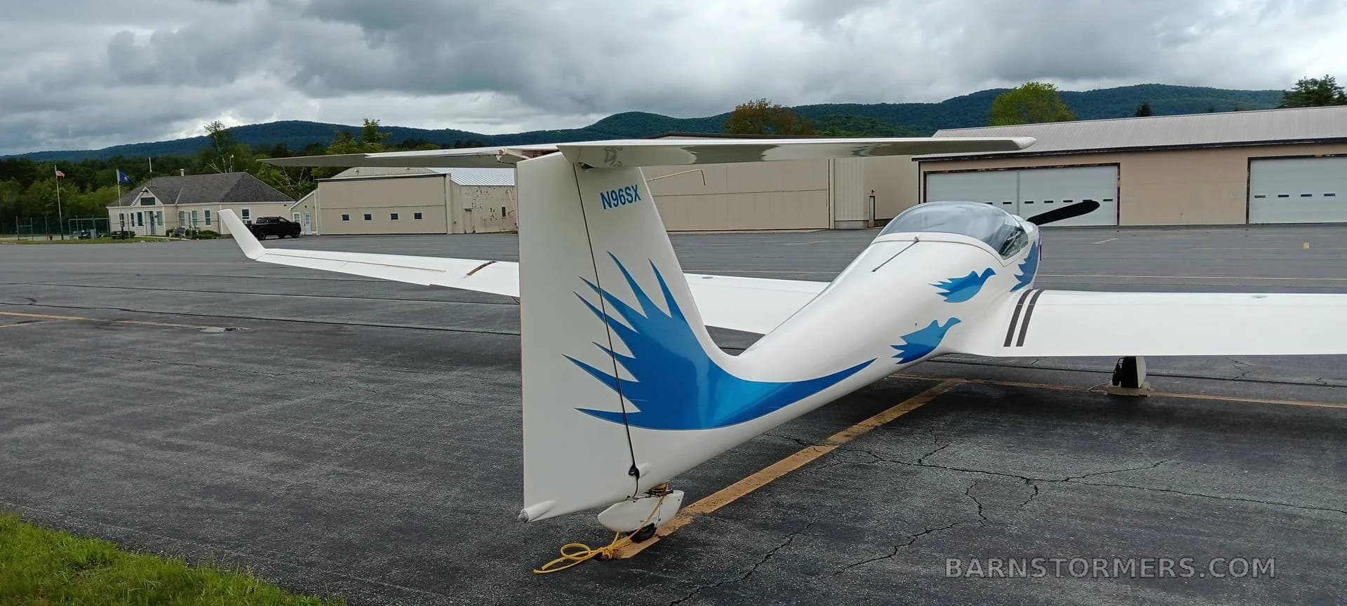 The tail and rear fuselage of a white motorglider with blue wave decals are seen from behind, parked on an airport tarmac with hangars and mountains in the background.