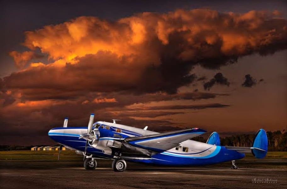 A blue and silver Beechcraft Model 18 parked on the tarmac with dramatic clouds in the background.
