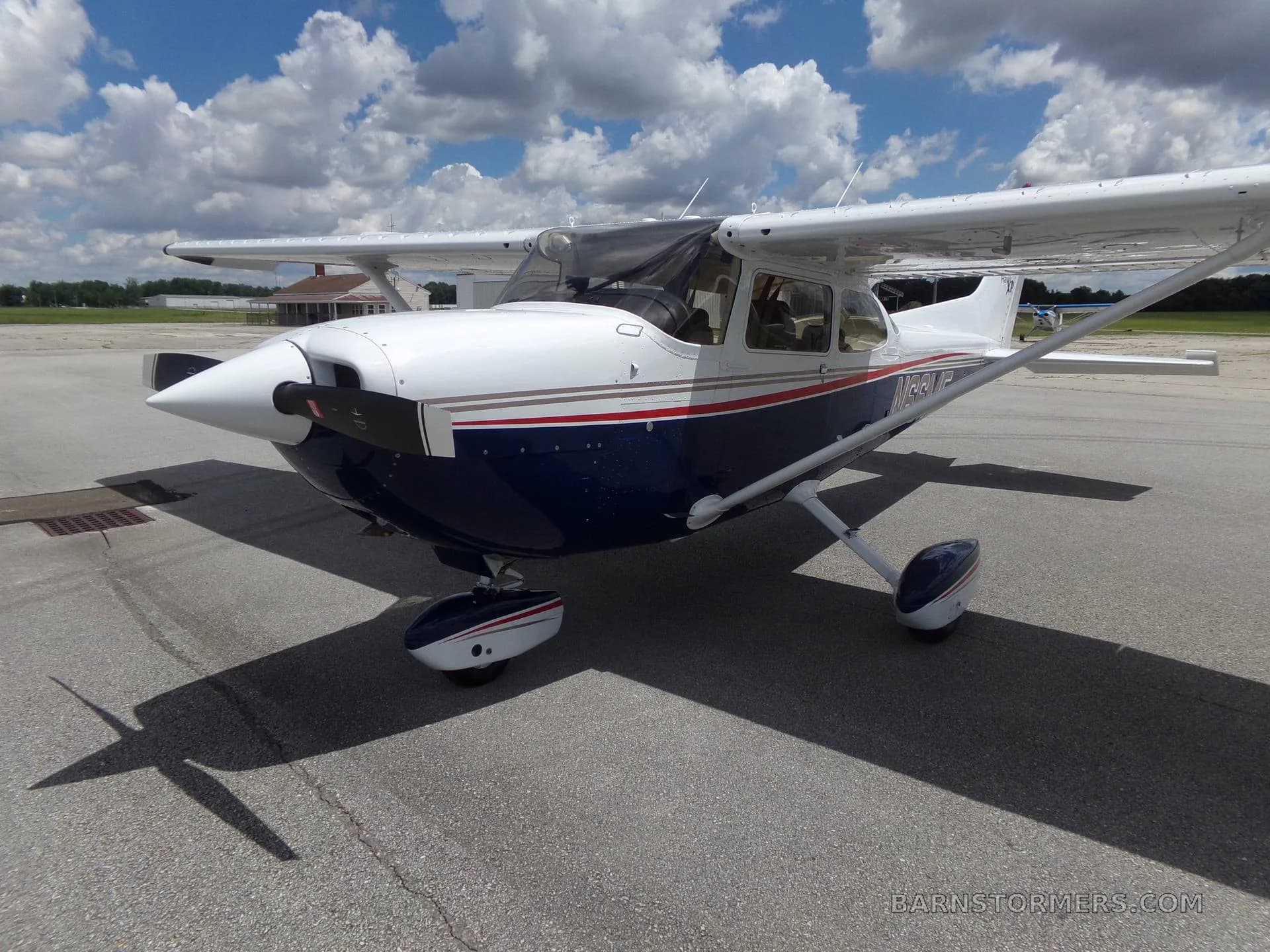 A white and blue Cessna 172 Hawk XP with red and gold stripes, seen from the front-right on an airfield under a partly cloudy sky.