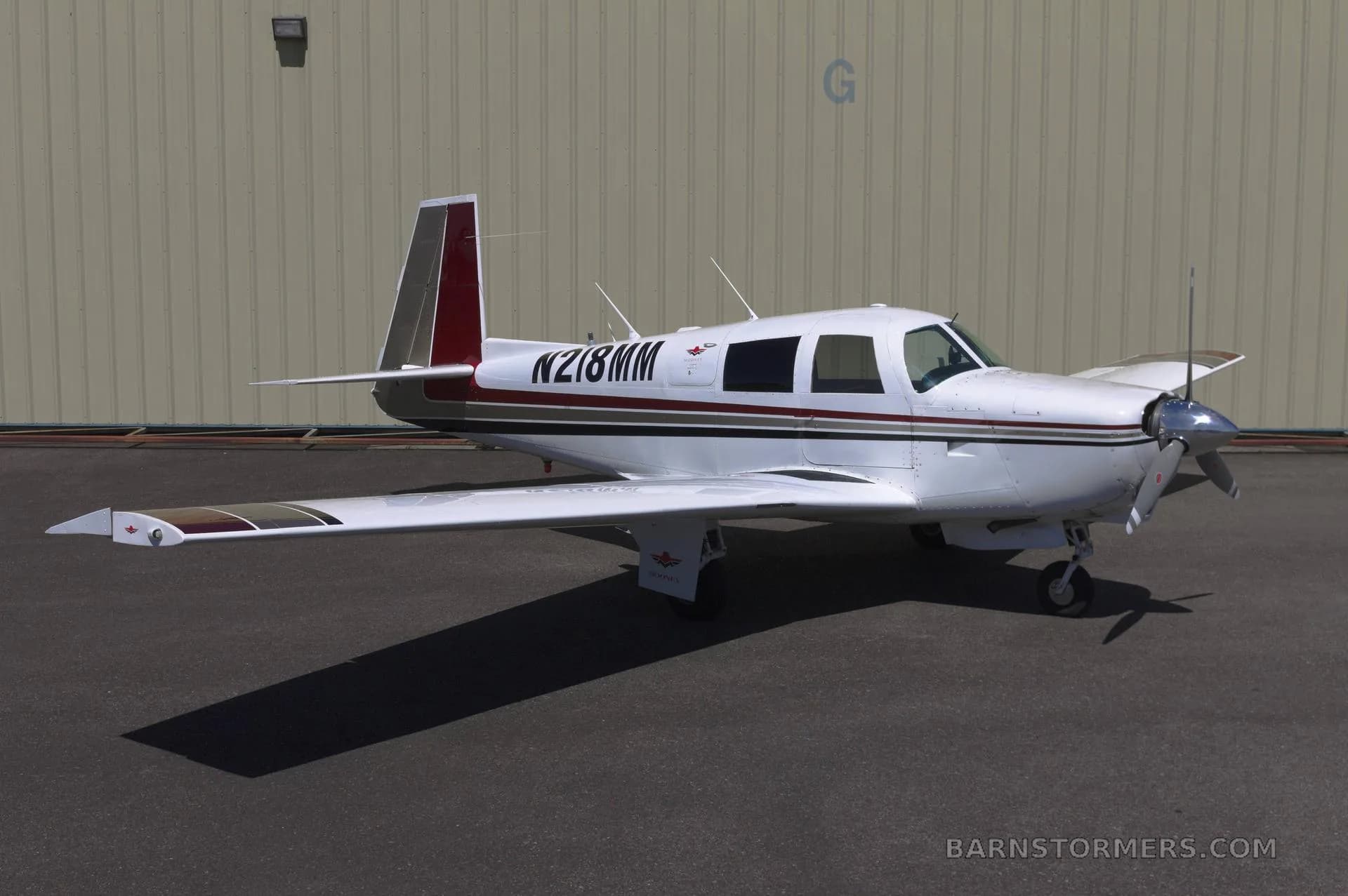 A white single-engine propeller aircraft with red and brown stripes on the tail and wings, parked on an asphalt surface in front of a light-colored hangar.