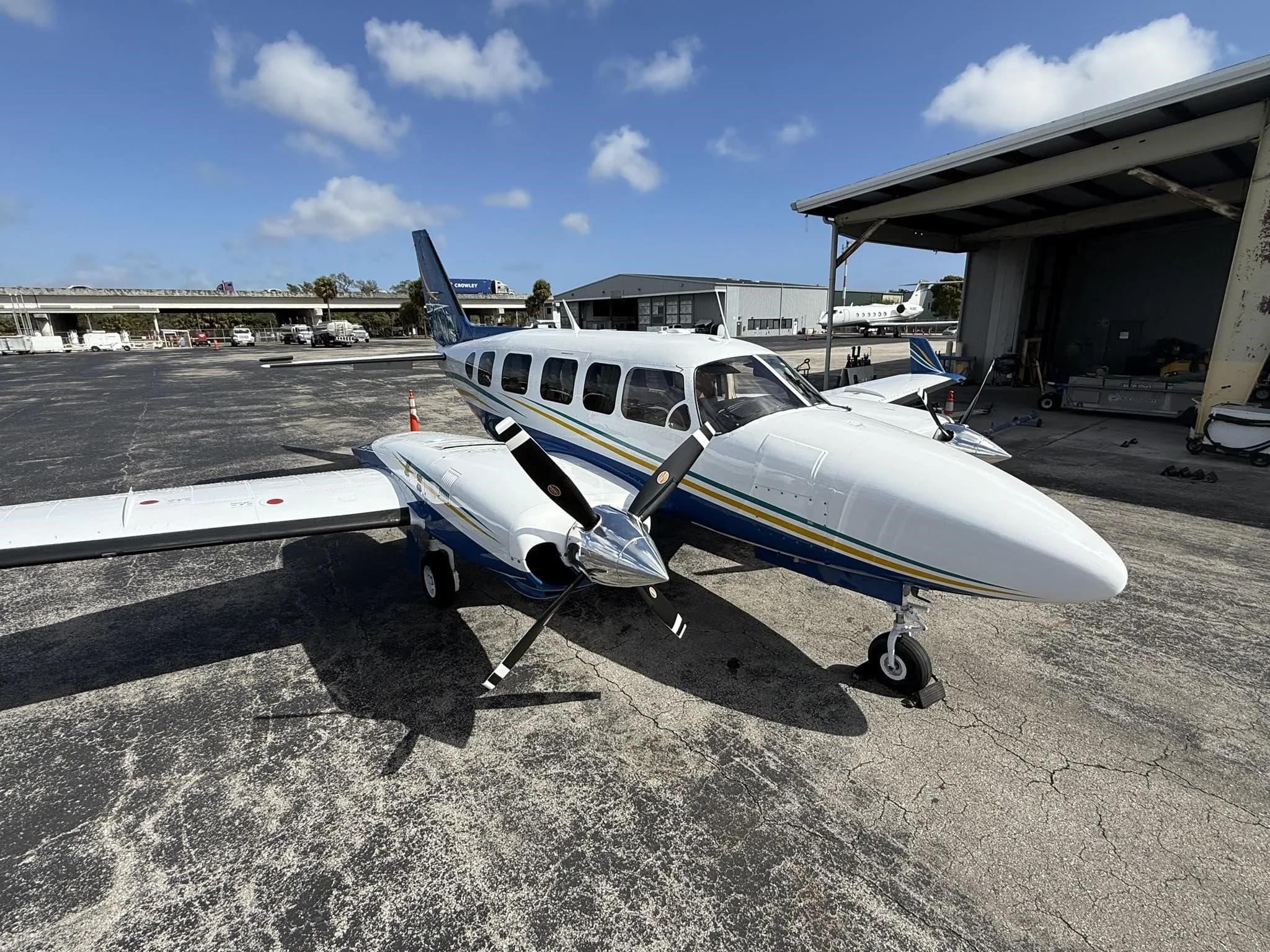 A white and blue twin-engine propeller aircraft with yellow and green stripes, parked on an airport tarmac next to a hangar.