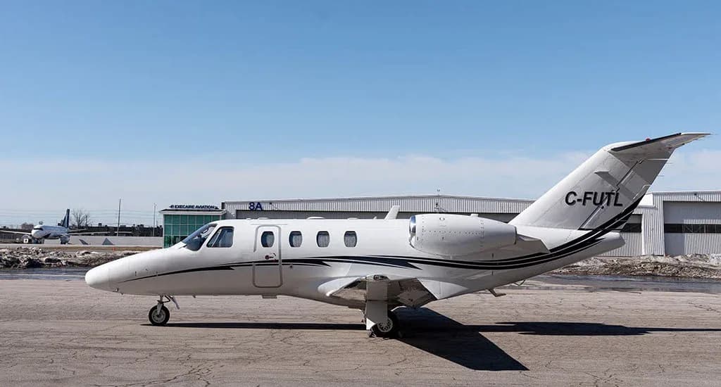 A white Cessna Citation business jet with black and grey stripes, registration C-FUTL, parked on a tarmac with hangars in the background.
