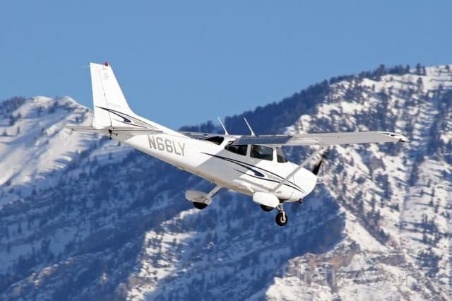A white Cessna 172 Skyhawk (N66LY) with blue stripes flying over snowy mountains.