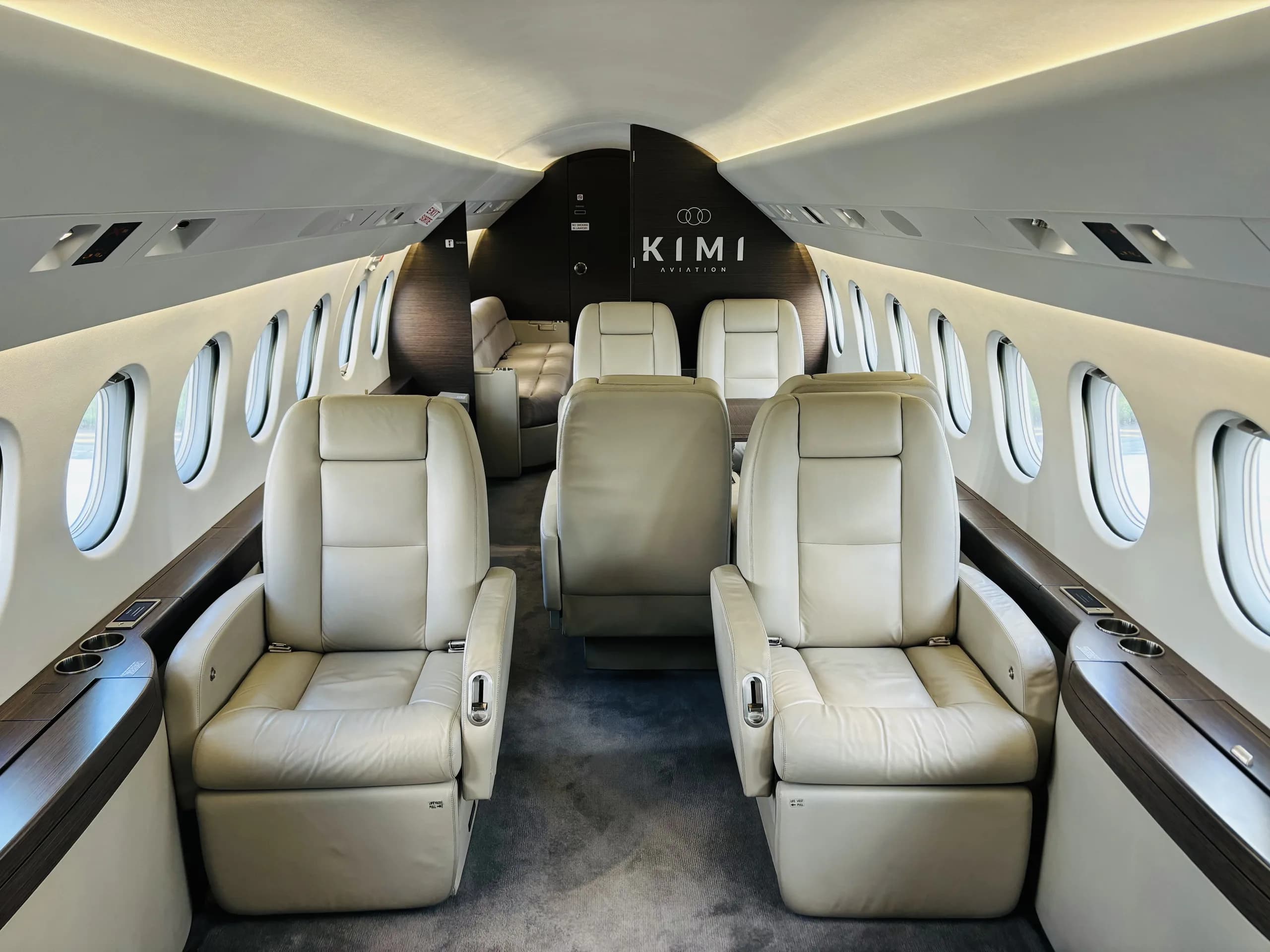 A wide view of a private jet's luxurious cabin interior, featuring multiple light-colored leather seats and dark wood tables.
