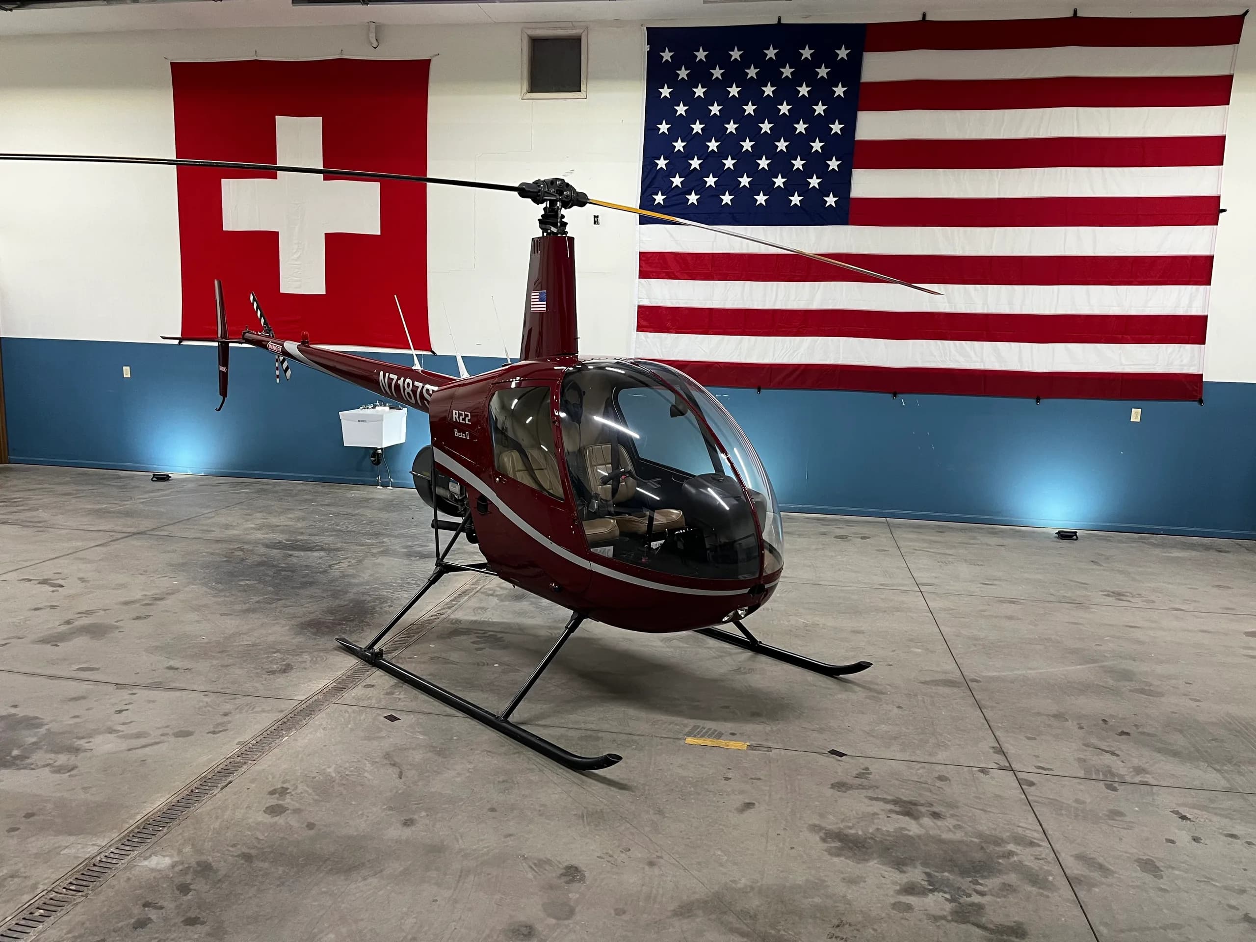 A red Robinson R22 helicopter, parked in a hangar with Swiss and American flags.