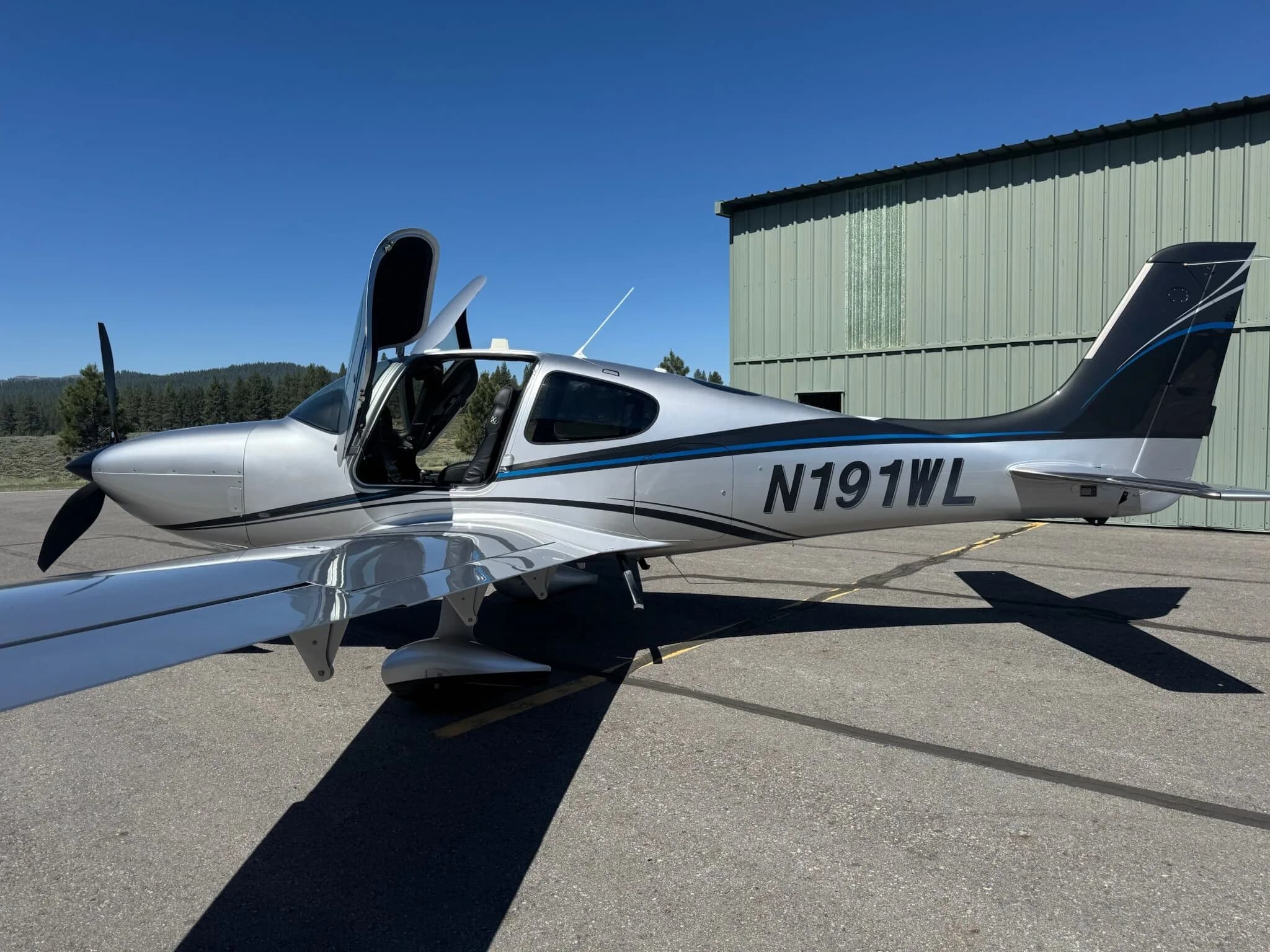 A side view of a silver and black Cirrus SR22T aircraft with N191WL, with the pilot's door open, parked next to a hangar.