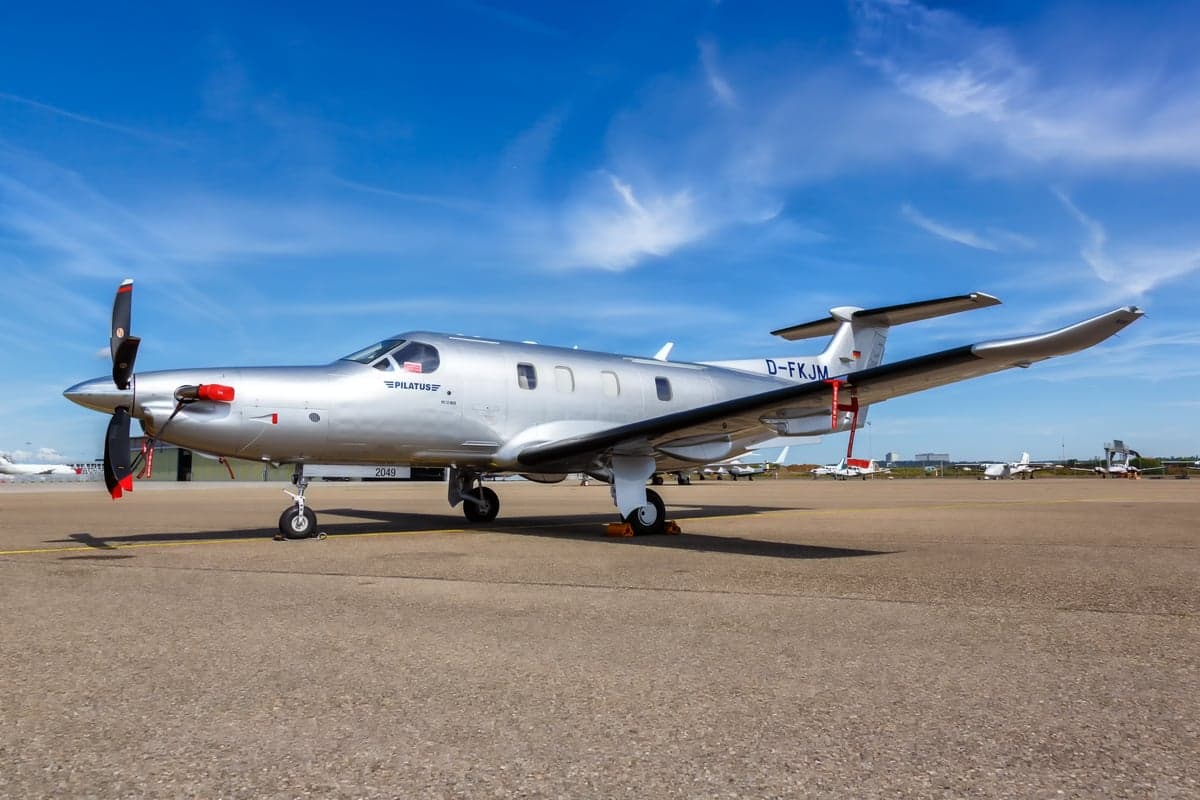 A silver Pilatus PC-12 is parked on the tarmac under a blue sky.