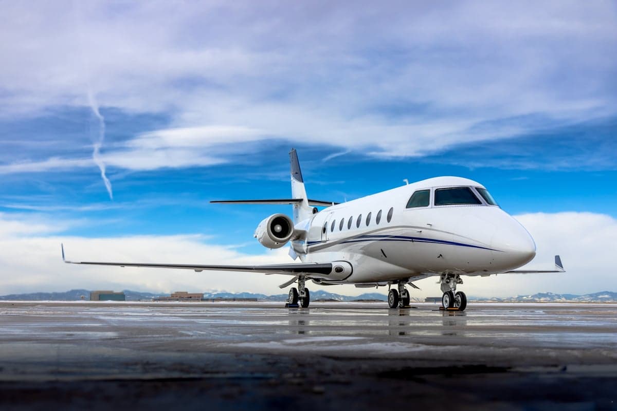 A white business jet with blue stripes parked on a wet tarmac under a partly cloudy sky.