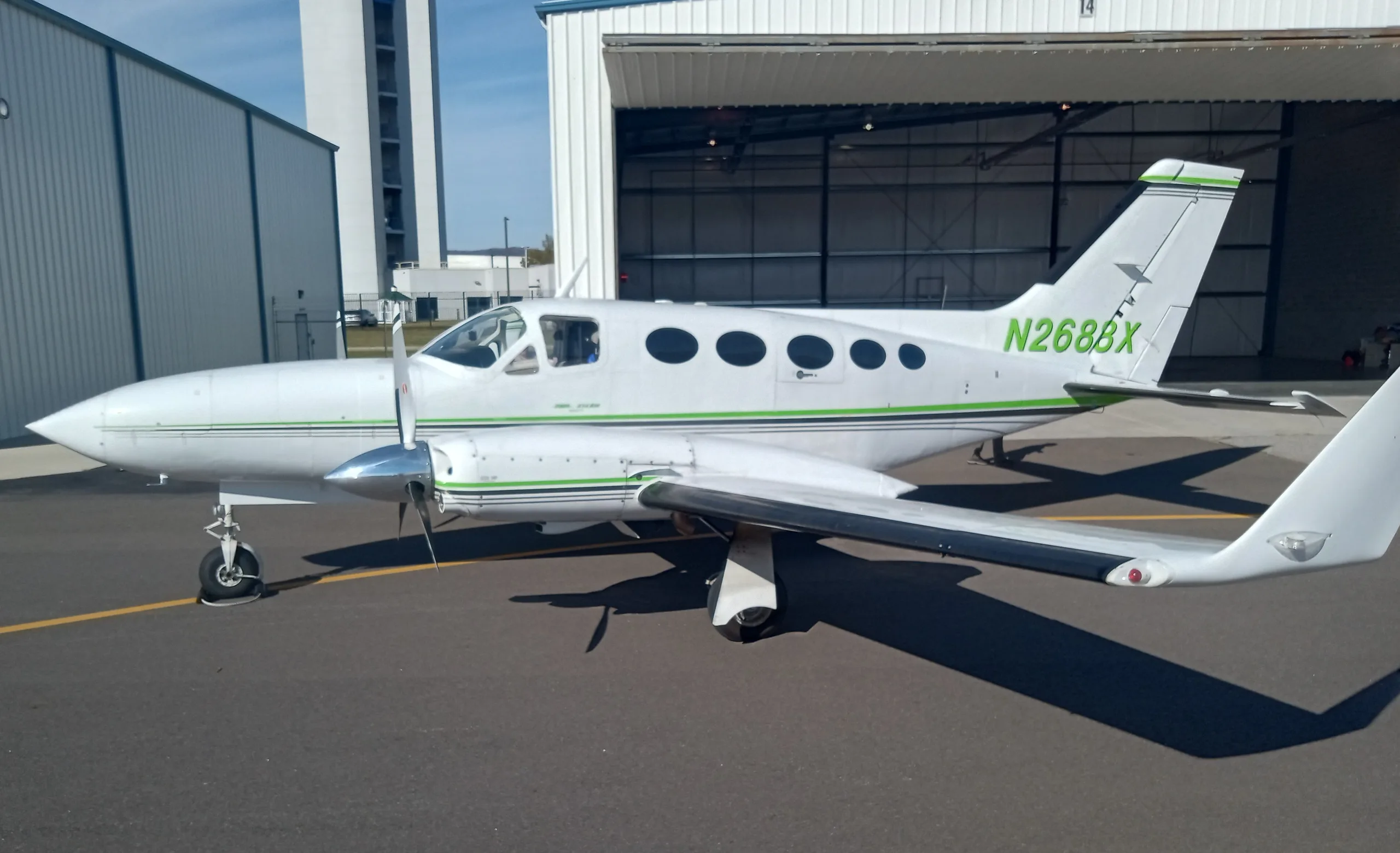 A white twin-engine propeller aircraft with green and black stripes, N2688X on the tail, parked on an asphalt ramp next to a hangar.
