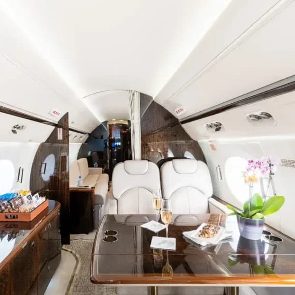 A wide view of the aircraft cabin interior showing a dining table with champagne and pastries, and a lounge area with a sofa in the background.