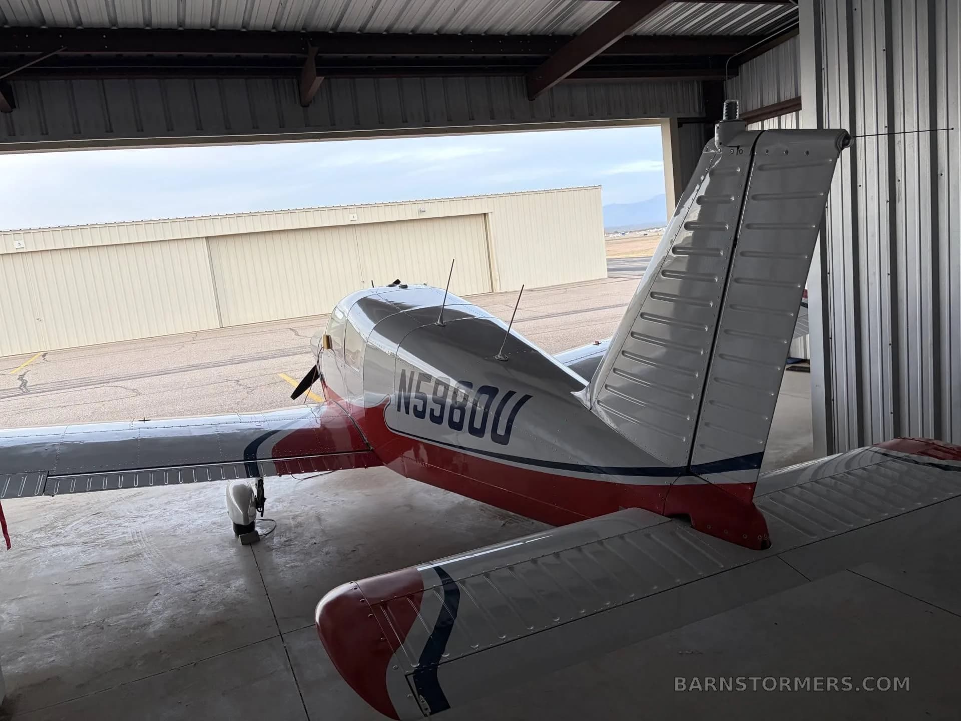 A rear view of the red, white, and blue single-engine propeller aircraft (N5980U) inside a hangar, looking out to the airfield.