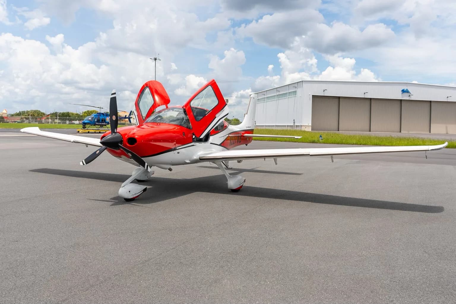 A red and white Cirrus SR22 aircraft with its gull-wing doors open, parked on an airfield with hangars in the background.