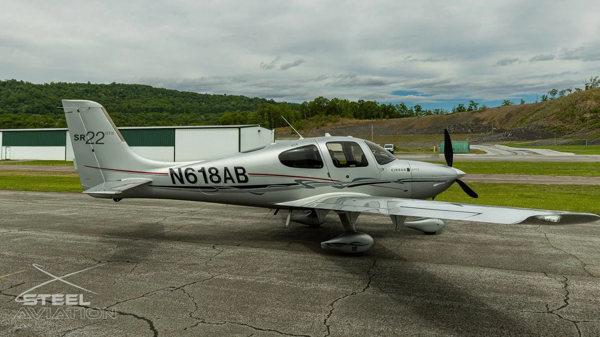 A silver Cirrus SR22 GTS aircraft with registration N618AB parked on an tarmac with buildings and hills in the background.