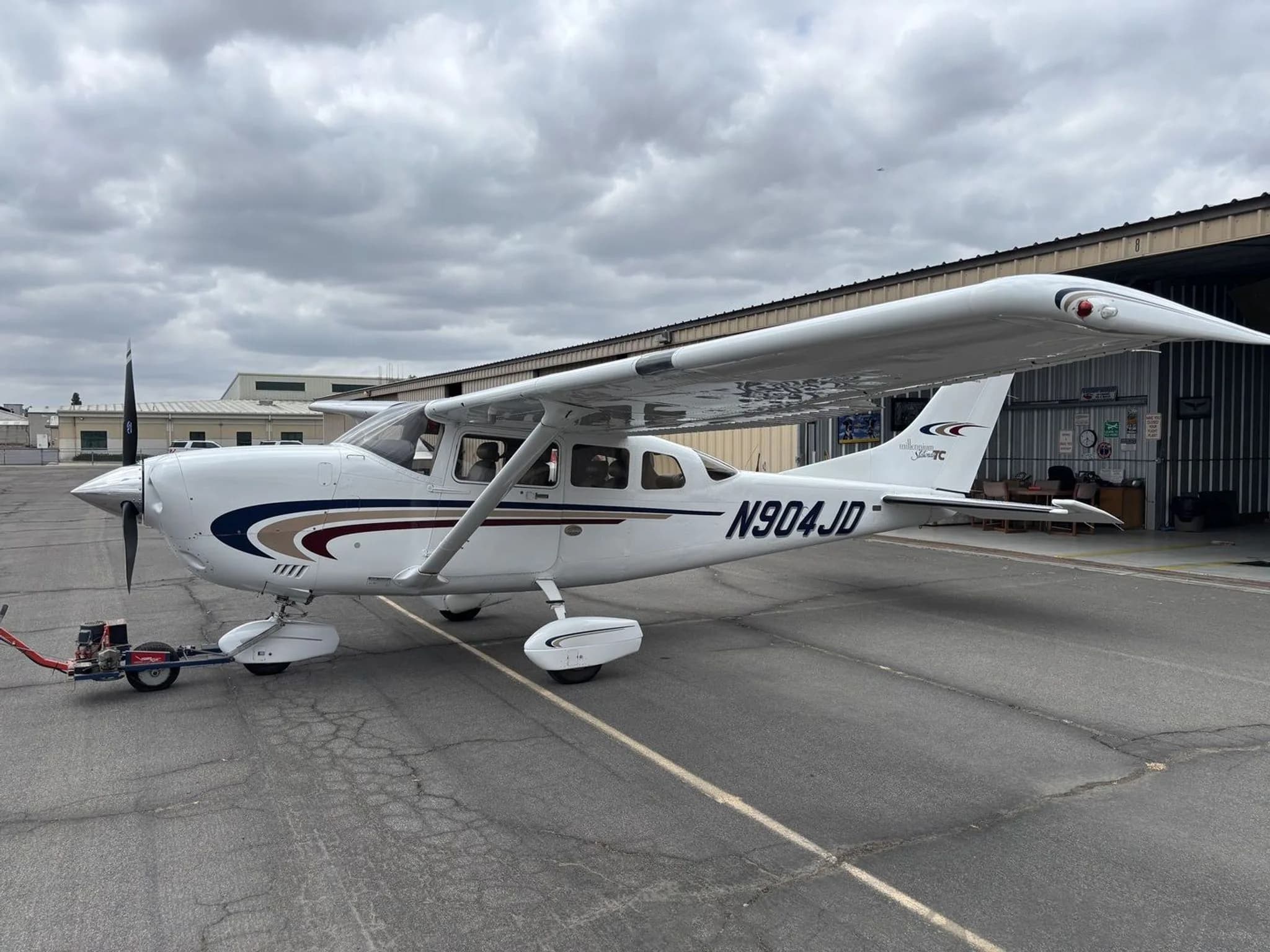 A white Cessna 182 Skylane TC aircraft with blue and maroon stripes, registration N904JD, parked on a tarmac with a hangar in the background.
