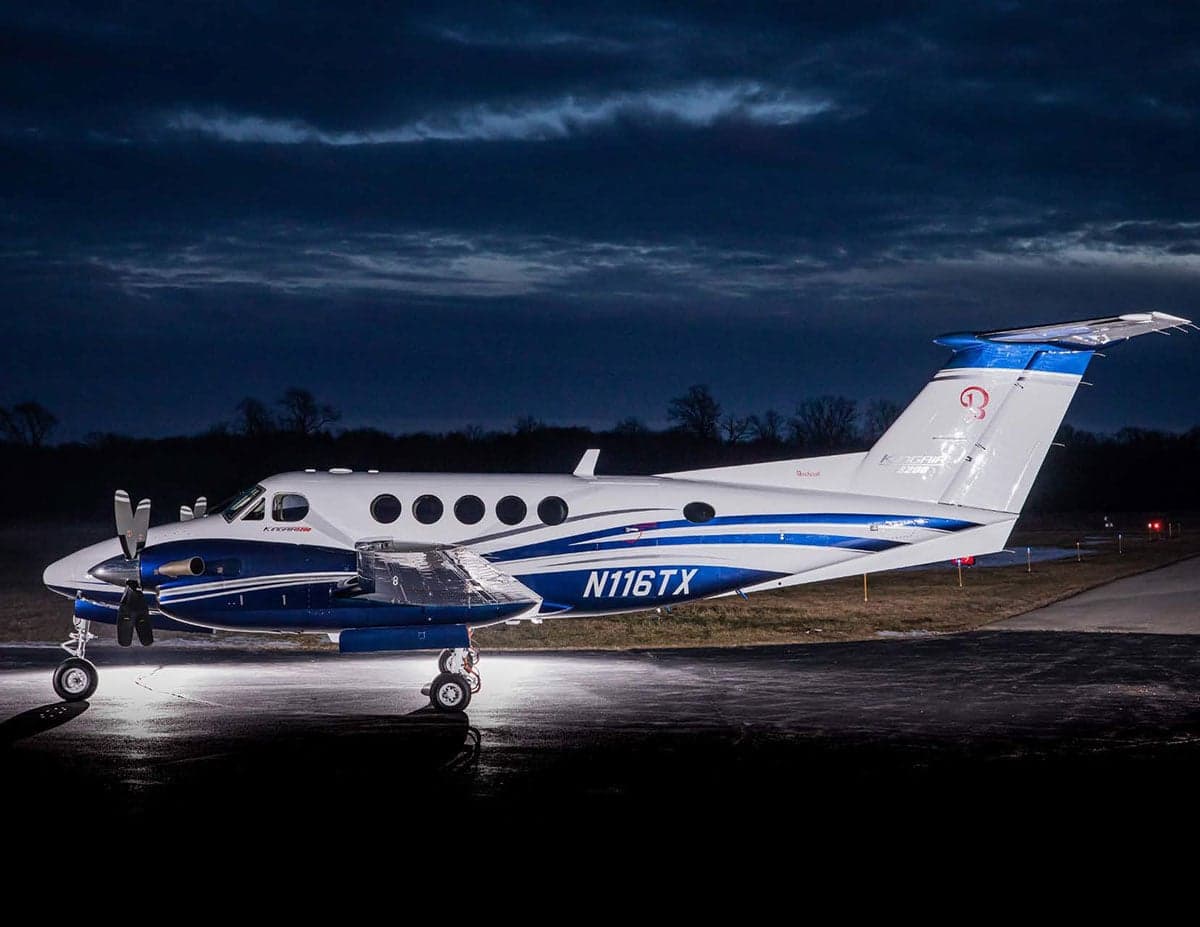 A white and blue Beechcraft King Air turboprop aircraft parked on an airfield at dusk.