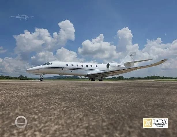A white Cessna Citation business jet is parked on an asphalt runway under a partly cloudy blue sky, with another aircraft faintly visible in the distance.
