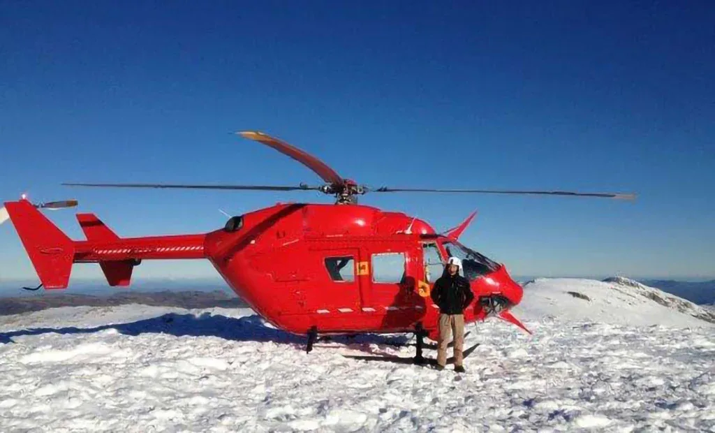 A red helicopter with skids parked on a snow-covered mountain peak with a person standing nearby.