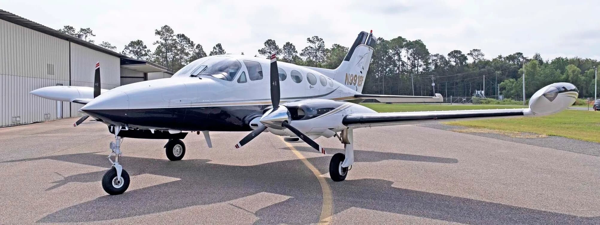 A front-quarter view of the white and dark blue Cessna 414 Chancellor, registration N991EM, parked in front of a hangar.
