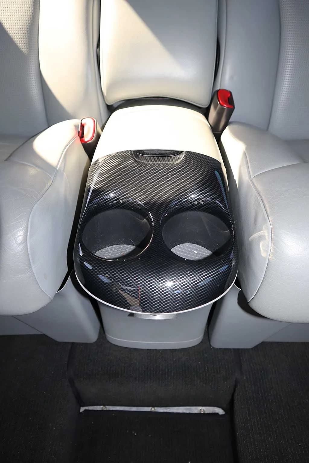 A close-up view of a black carbon fiber-patterned center console with two cup holders between two light gray seats.