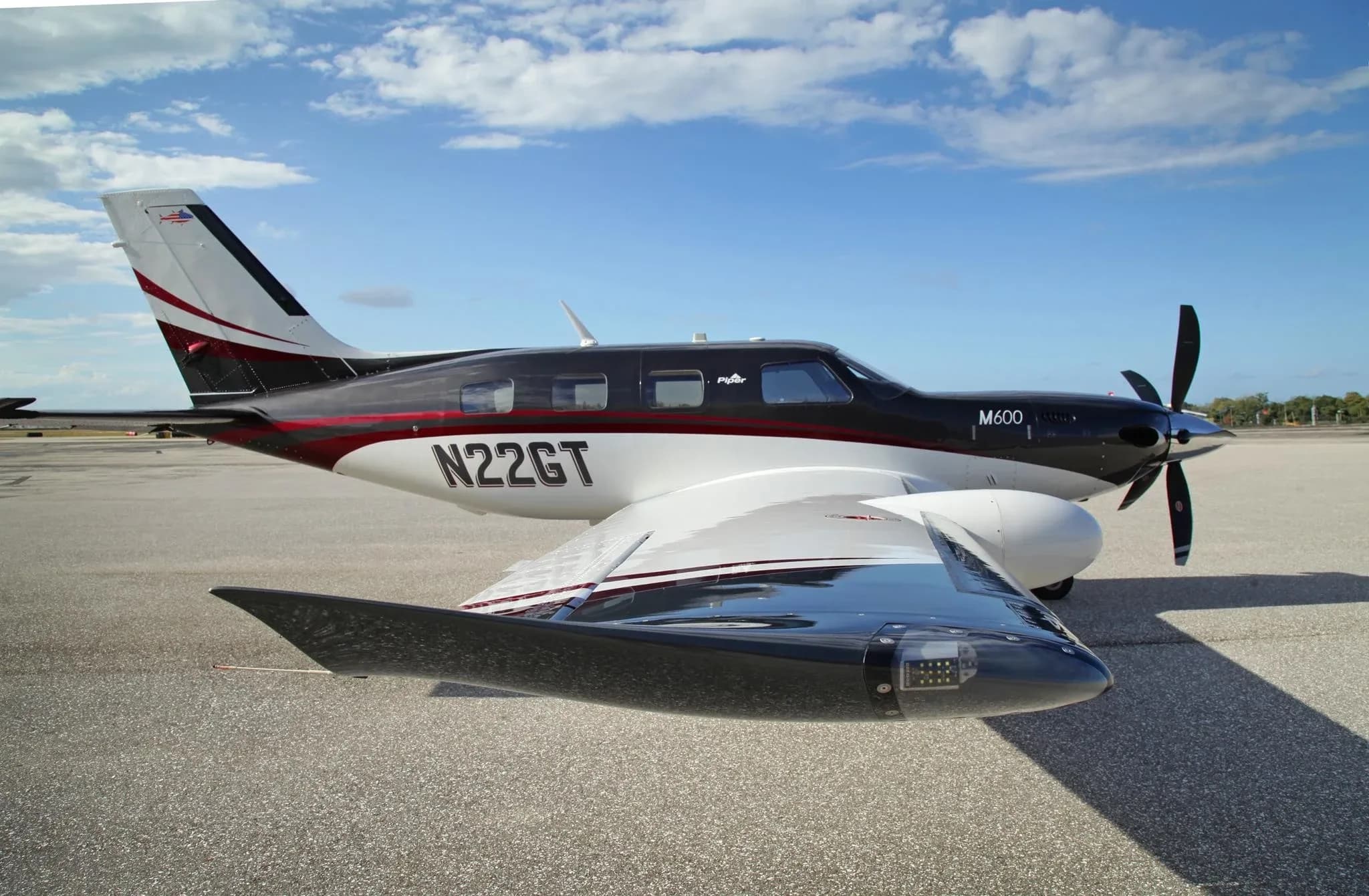 A Piper M600 aircraft with N22GT registration, featuring a black, white, and red paint scheme, parked on an airfield under a blue sky.