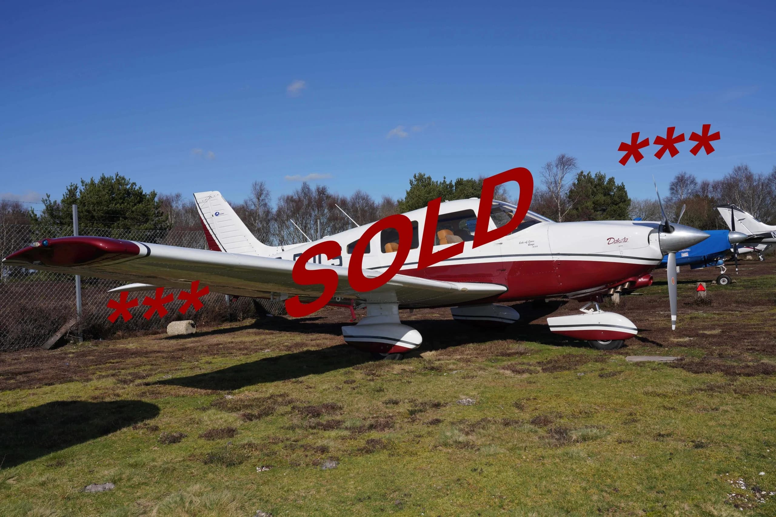 A white and red Piper Dakota aircraft is parked on a grassy field under a clear blue sky, with the word "SOLD" overlaid in red.