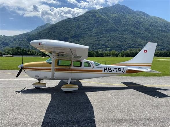 A Cessna 210 Centurion aircraft with a white and gold paint scheme is parked on the tarmac with mountains in the background.