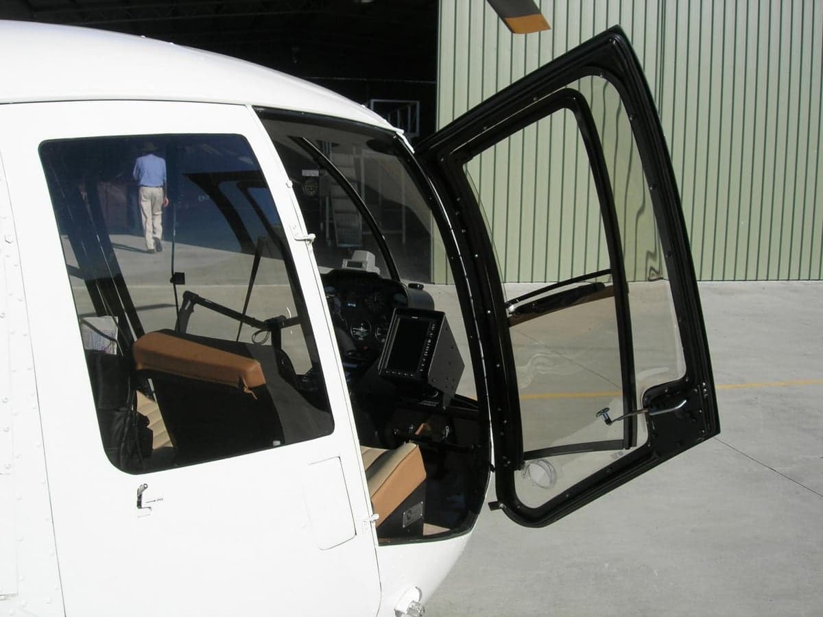A side view of a white helicopter with its door open, revealing the cockpit and a person walking in the background near a hangar.