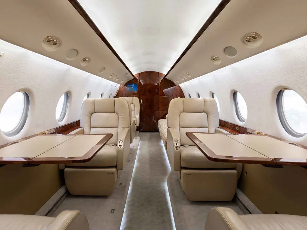 The cabin interior of a Gulfstream G450 showing beige leather seats with fold-out tables deployed.