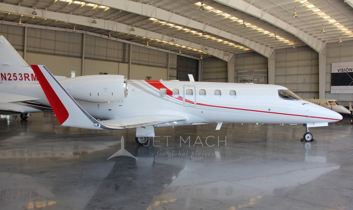 A white Learjet 75 with red and grey stripes, registration N253RM, parked inside a hangar.