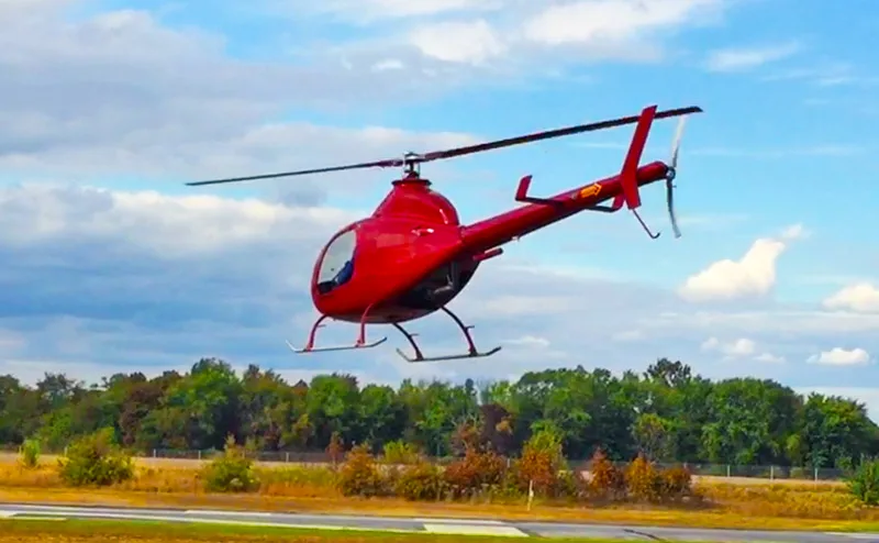 A red RotorWay Exec helicopter is seen in flight from a rear-side view against a cloudy sky.