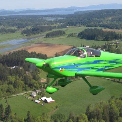 A green aircraft with blue accents flying over a rural landscape.
