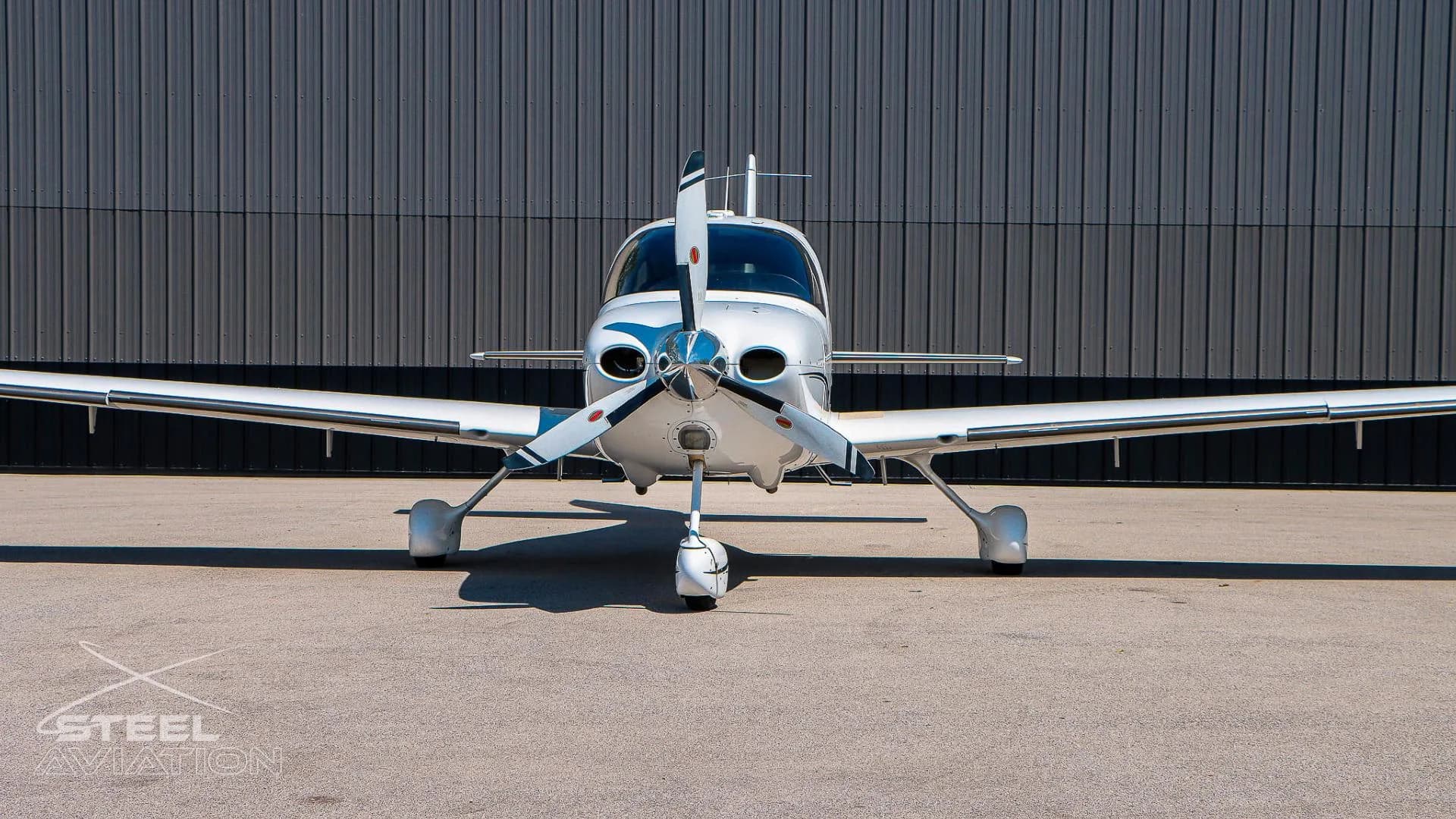 Close-up front view of a white Cirrus SR22 aircraft, showing its propeller and nose, in front of a grey hangar.