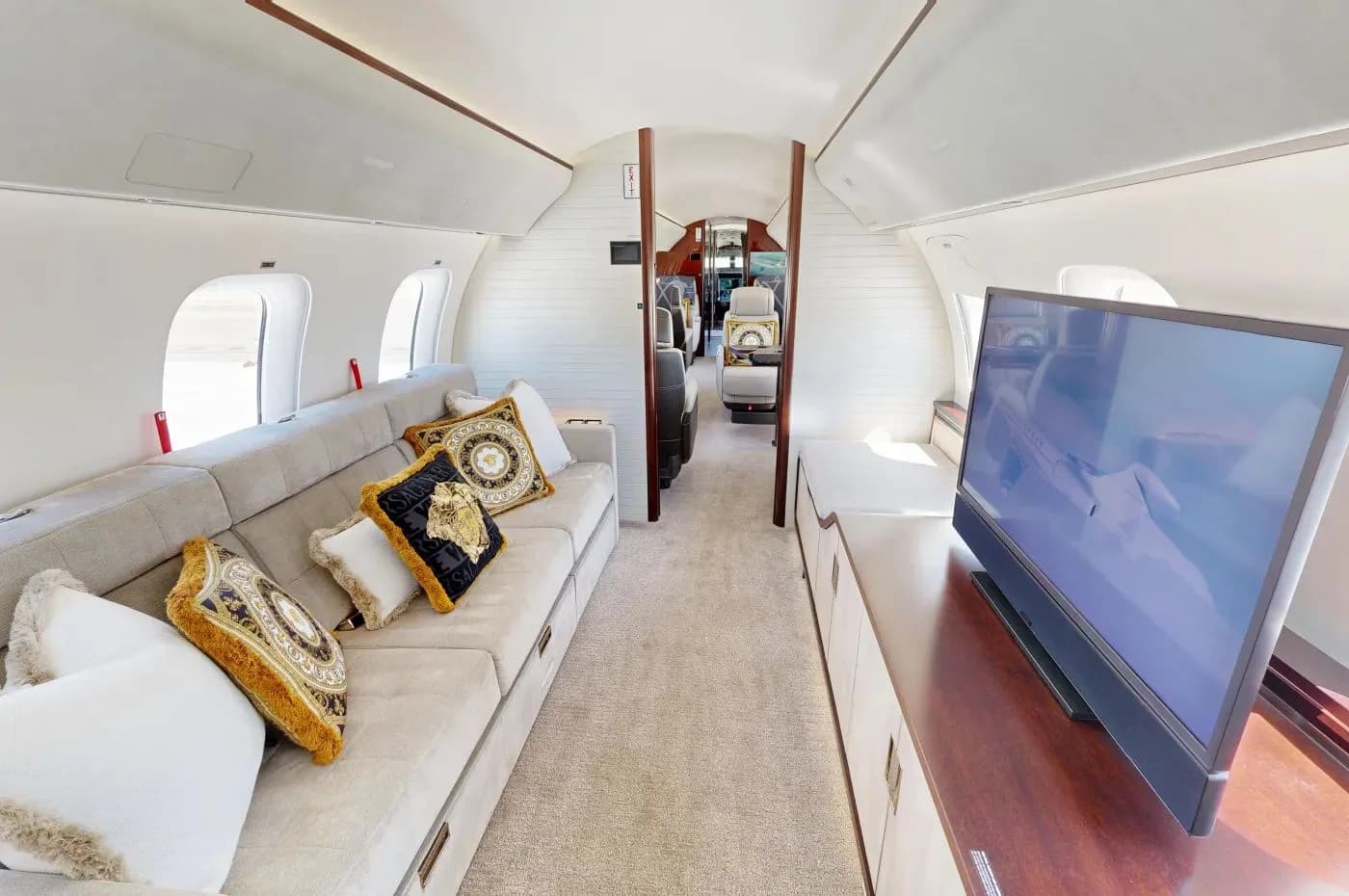 The main cabin of a private jet featuring a long grey sofa with decorative pillows and a large television screen.