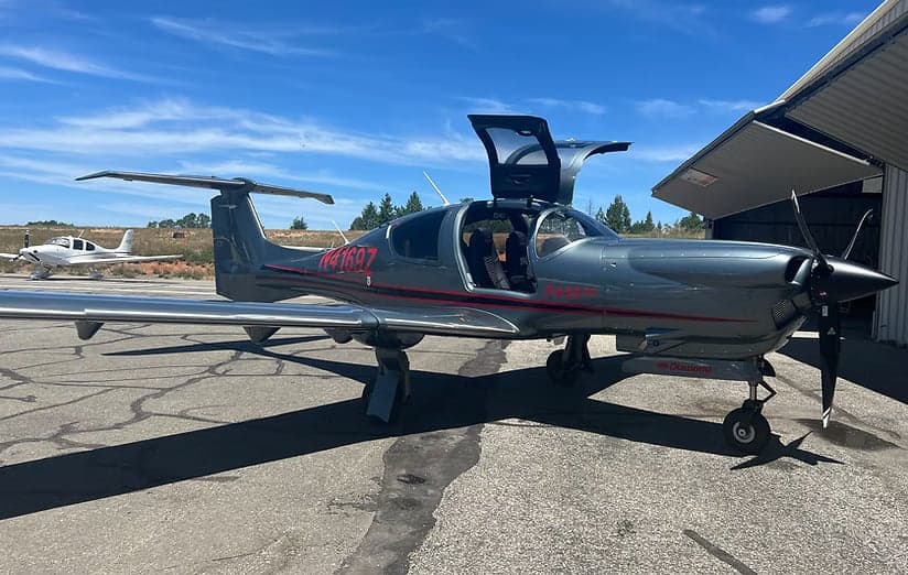 A gray Diamond DA42-VI aircraft is parked on the tarmac with its gull-wing doors open.