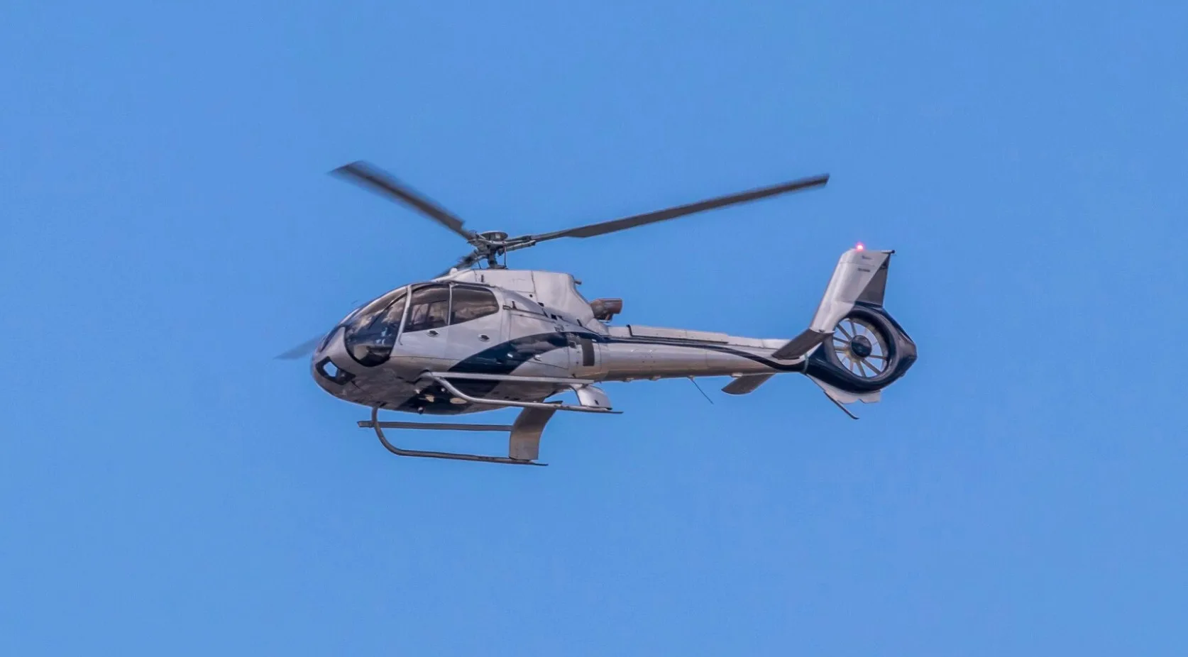 A silver and black Eurocopter EC130 B4 helicopter in flight against a blue sky.