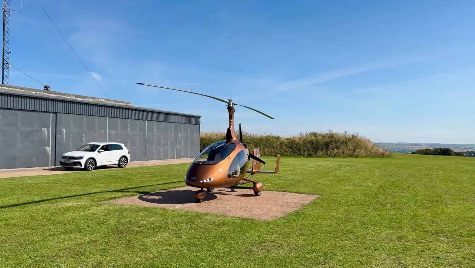 A brown gyrocopter parked on a concrete pad in a grassy field with a white SUV and hangar in the background.
