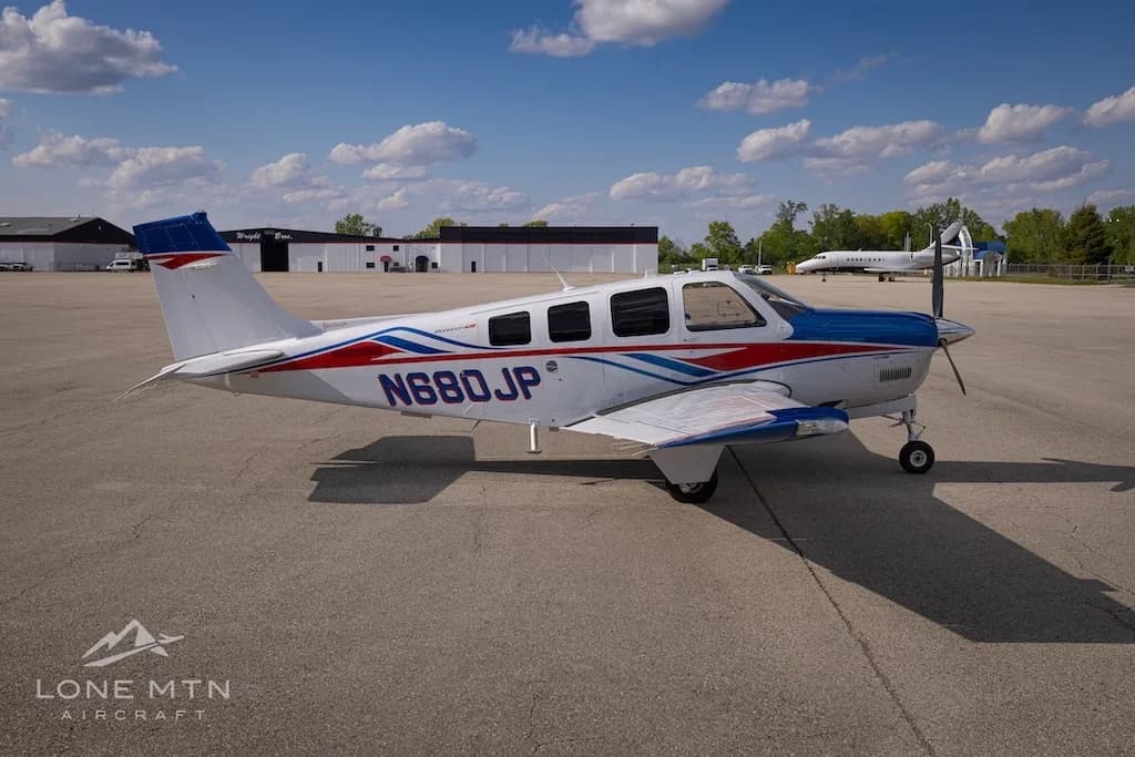 A side view of a white, blue, and red Beechcraft Bonanza N680JP parked on an airfield with other buildings in the background.