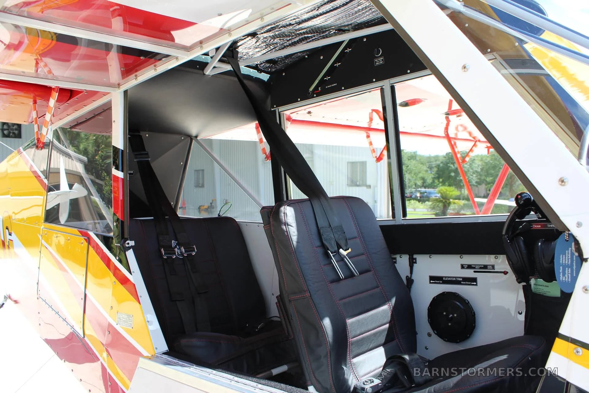 The interior of a light aircraft cabin, showing two black leather seats with red stitching and harnesses, looking towards the rear.