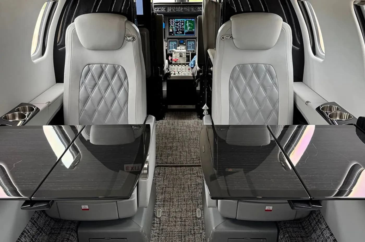 The forward cabin interior of a business jet with light grey quilted leather seats and dark polished tables, looking towards the cockpit.