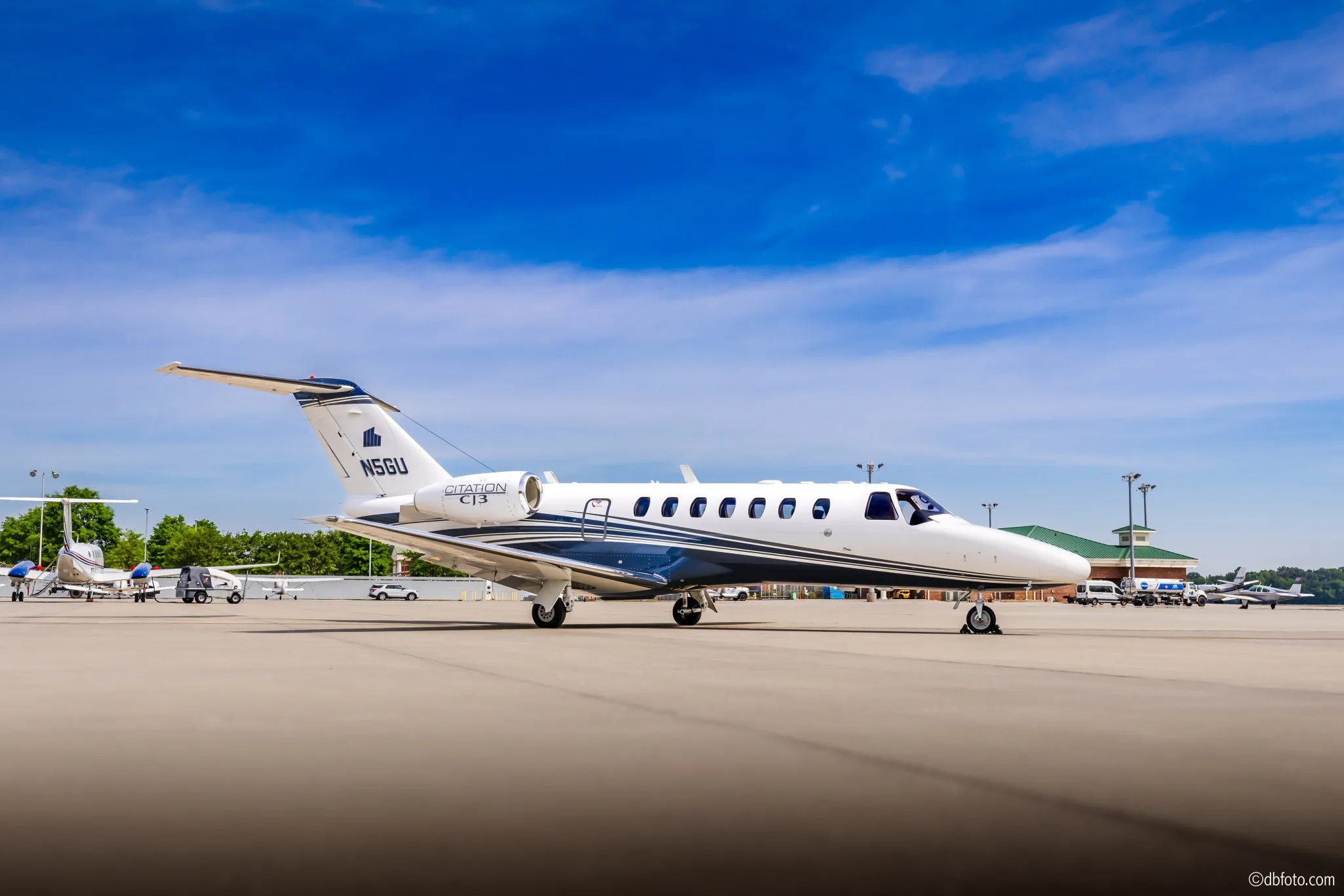 A white Cessna Citation CJ3 business jet with blue and grey stripes, registration N5GU, parked on an airport tarmac with other aircraft in the background.