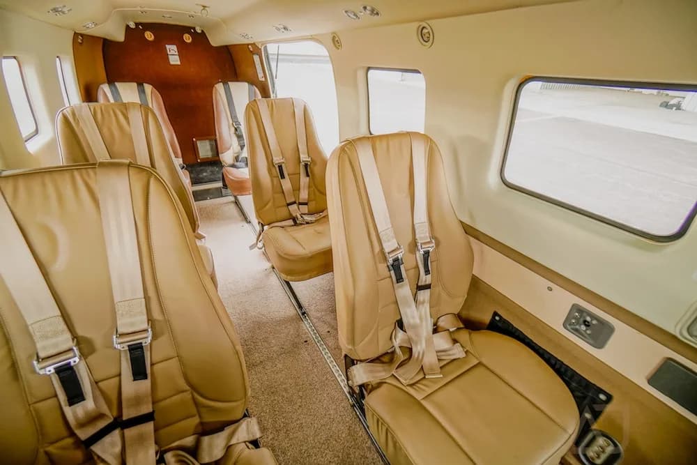 The aircraft interior cabin with tan seats and shoulder harnesses, showing a window view of the tarmac.