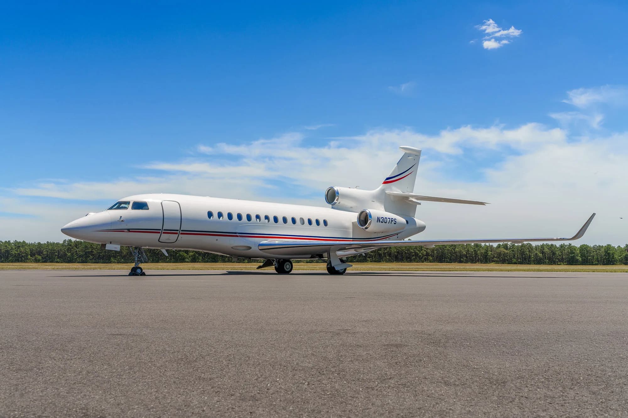 A white Dassault Falcon business jet with red and blue stripes is parked on an asphalt tarmac under a blue sky.