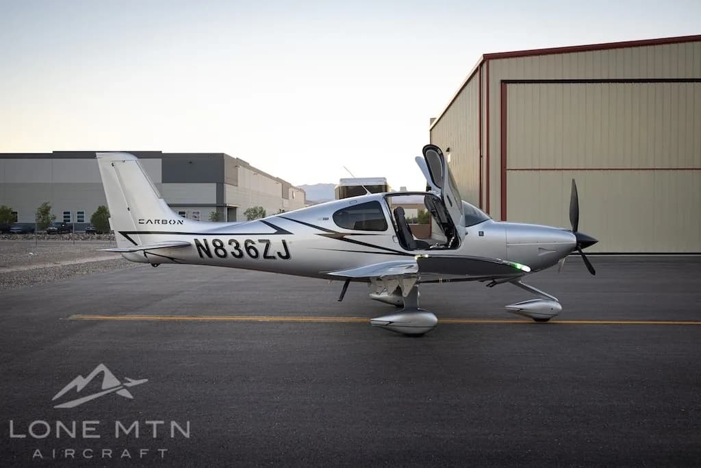 A silver Cirrus SR22T aircraft with registration N836ZJ is parked on an asphalt ramp with its doors open, with industrial buildings in the background.