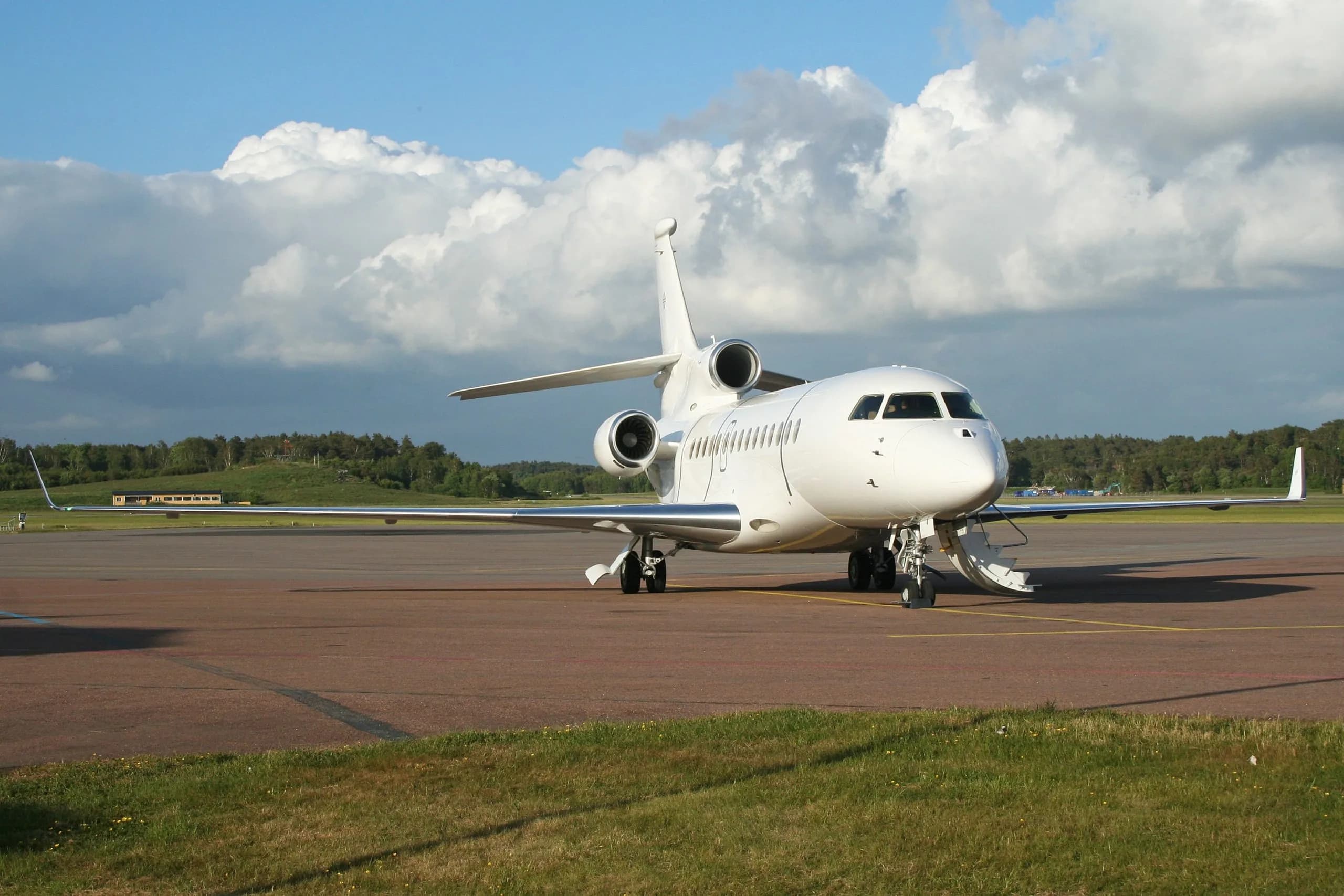 A white Dassault Falcon 900 trijet business jet parked on an airport apron under a partly cloudy sky.