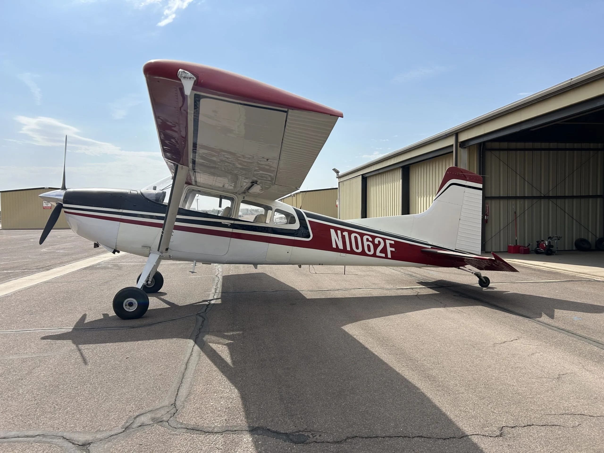 A white, black, and red Cessna 180 aircraft (N1062F) parked on an airport tarmac with its wing slightly raised.