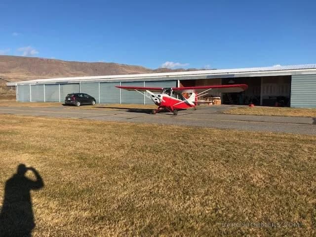 A red and white light aircraft parked on an asphalt strip next to a hangar under a clear blue sky.