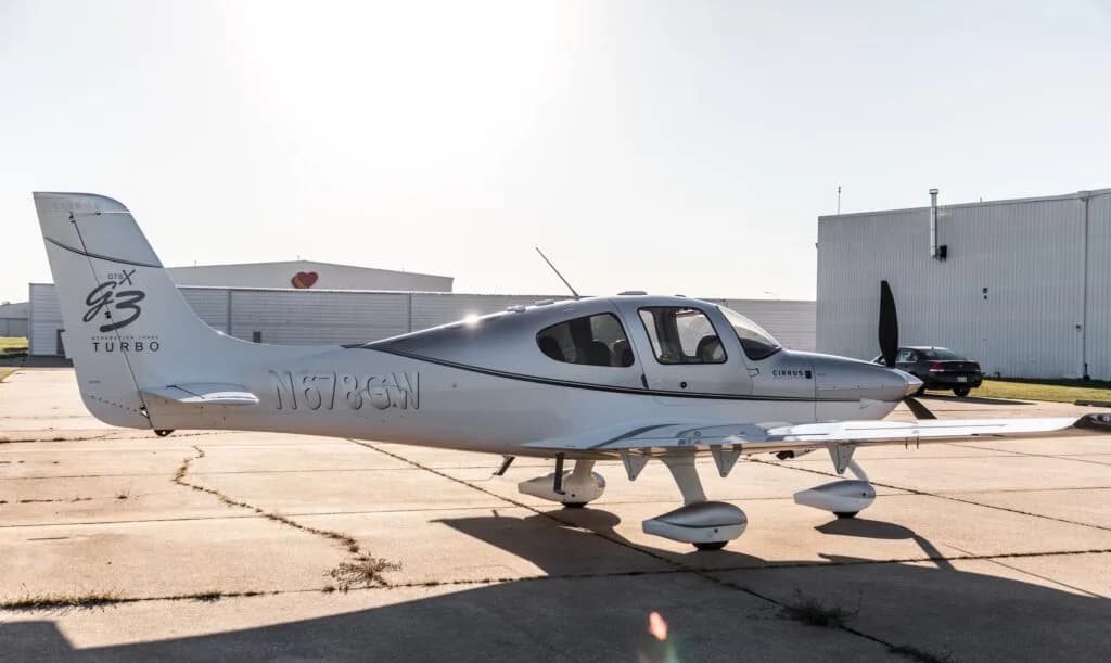 A side view of a silver and white Cirrus SR22 G3 Turbo aircraft (N678GW) parked on an airfield with hangars in the background.