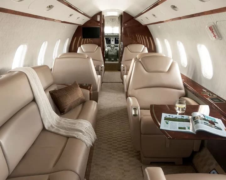 The luxurious cabin interior of a business jet features beige leather seats, a sofa, and dark wood accents, with the cockpit visible in the background.