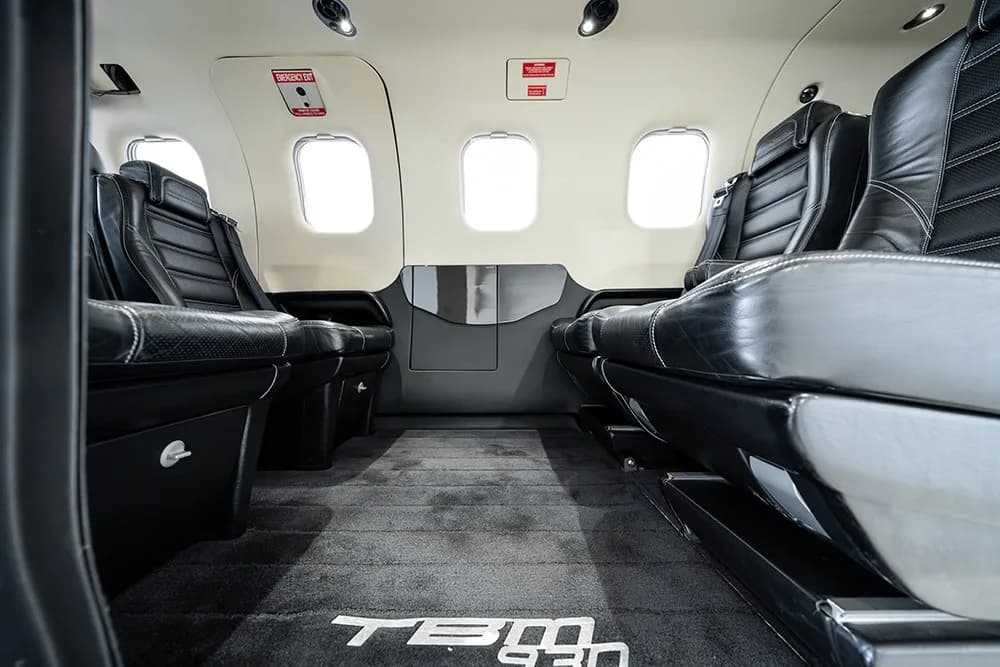 A view looking aft in the TBM 930 cabin, showing black leather seats and the "TBM 930" logo on the carpet.