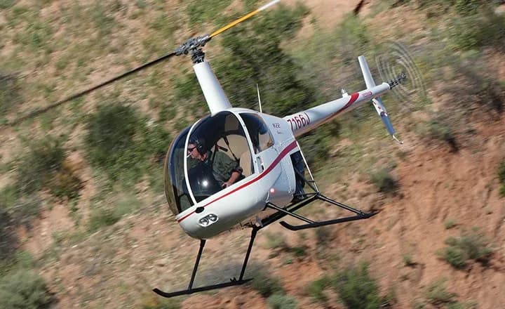 A white Robinson R22 Beta helicopter with red stripes flying over a hilly, vegetated landscape.